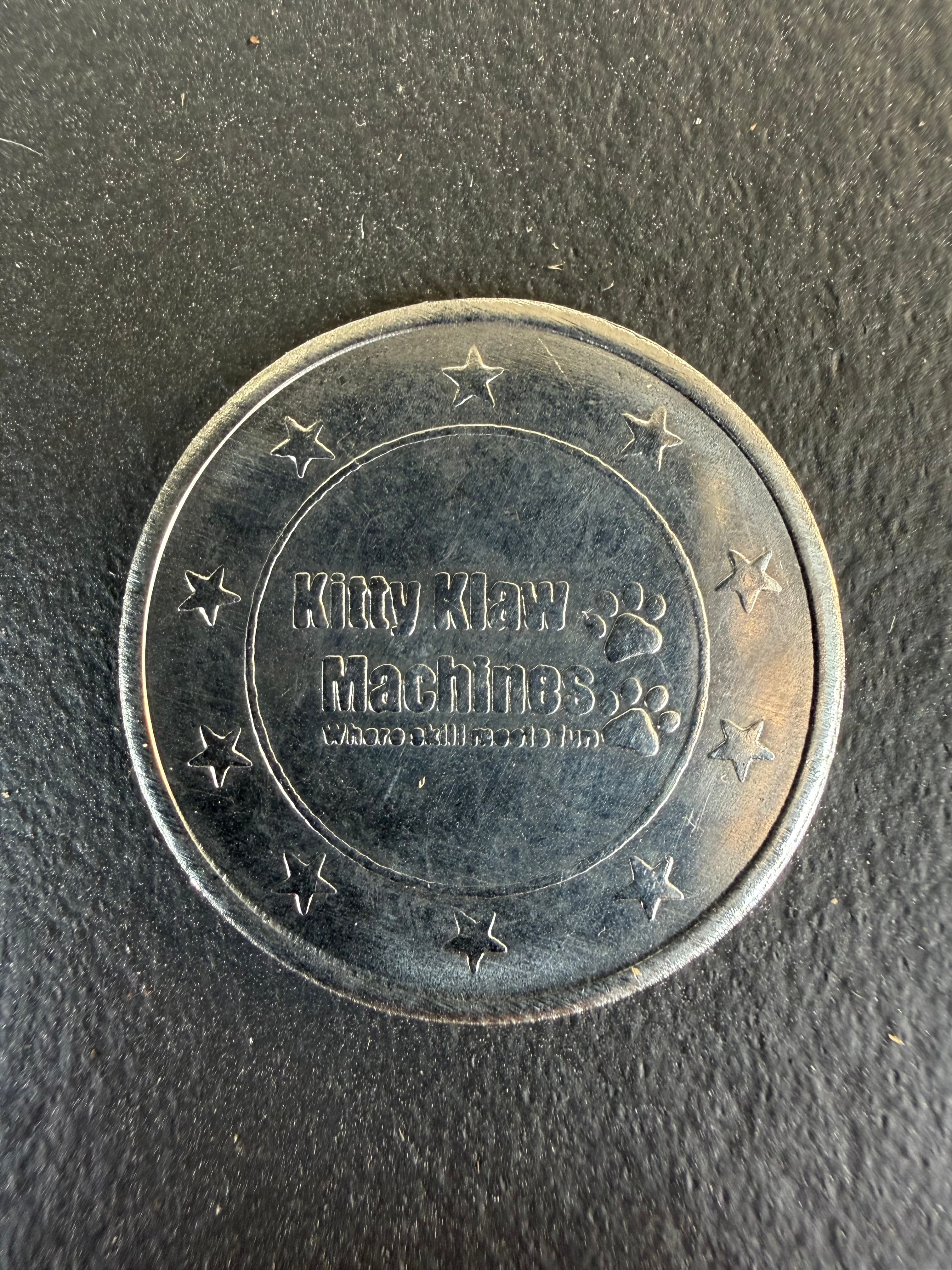 Silver Kitty Klaw Machines arcade token with stars and paw prints on dark textured surface.
