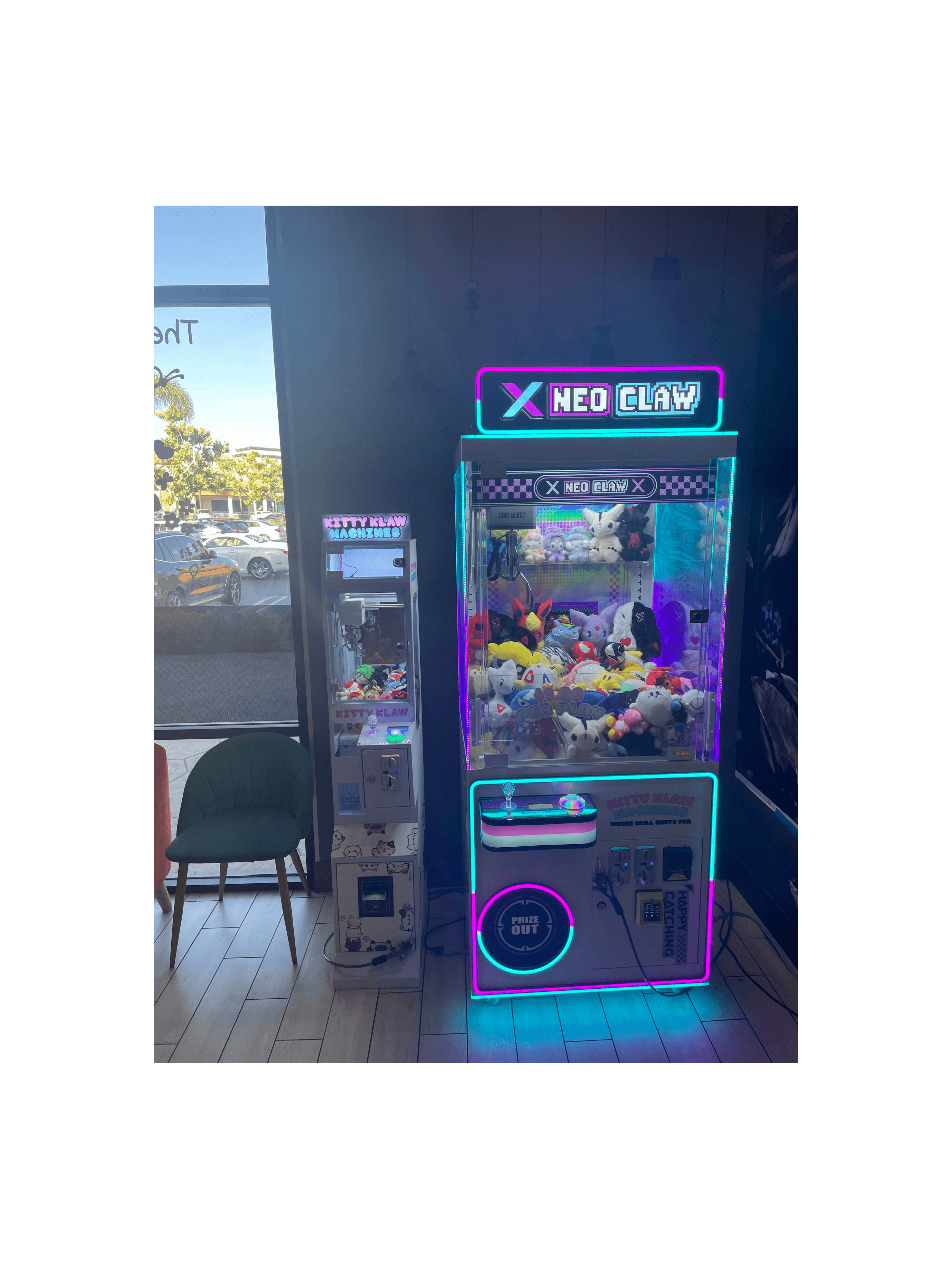 Two neon-lit claw machines filled with plush toys stand indoors next to a green chair.